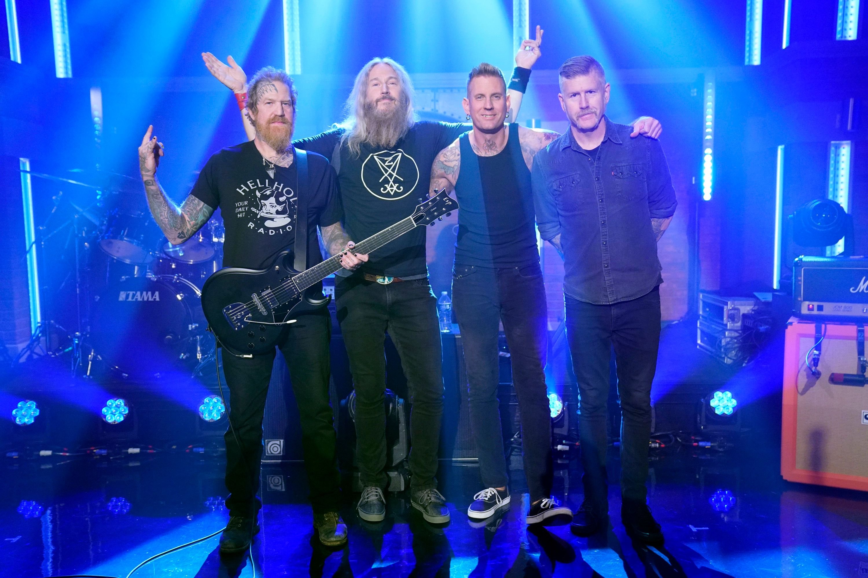 Four men pose together on a blue-lit stage with arms raised, one holding a black electric guitar, with drum kit and amplifiers visible behind them.