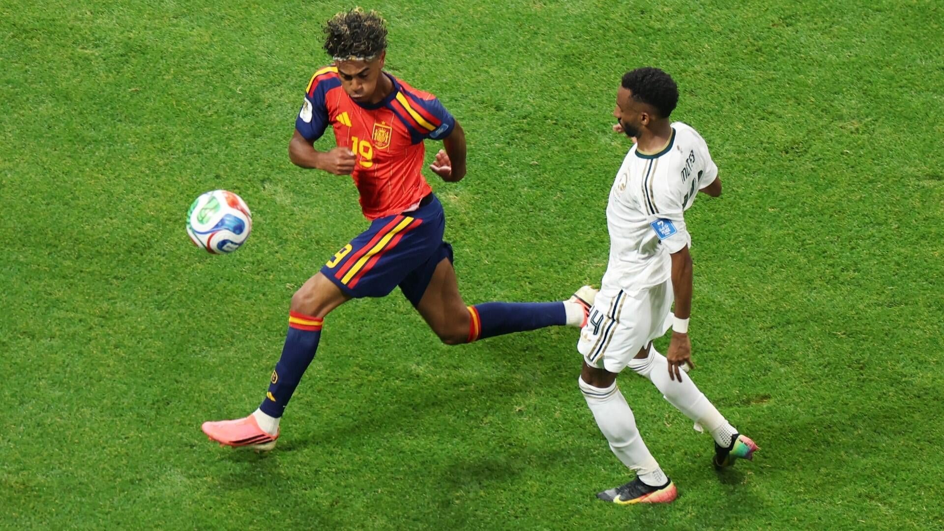 A soccer player in a red jersey numbered 19 runs with a ball on a grass field while an opponent in white pursues closely behind.