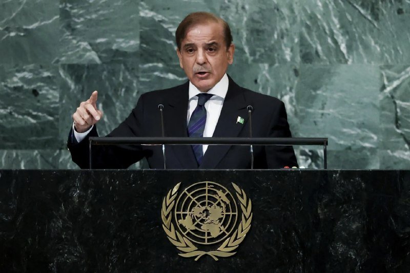 A man in a dark suit and striped tie speaks at a podium bearing the United Nations emblem, gesturing with one hand against a green marble backdrop.