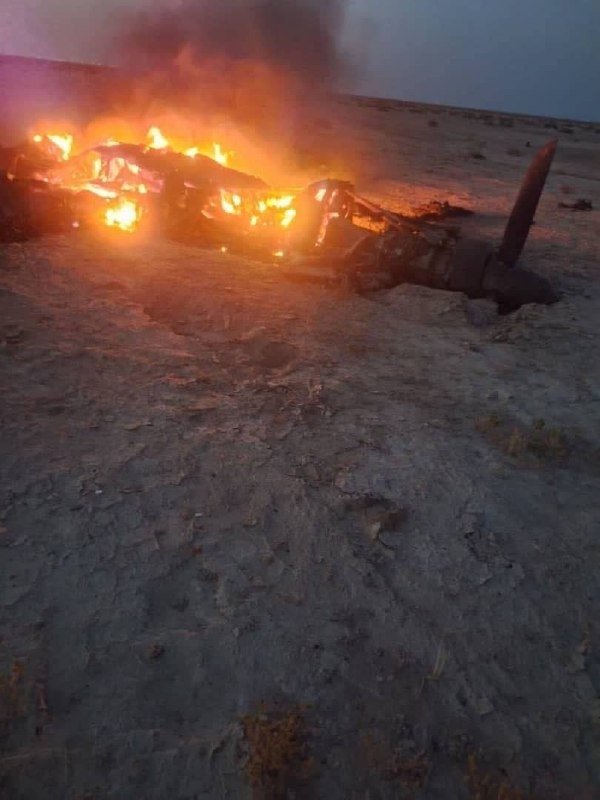 A burning aircraft wreckage with a visible propeller rests on barren dirt terrain as flames and smoke rise from the fuselage.