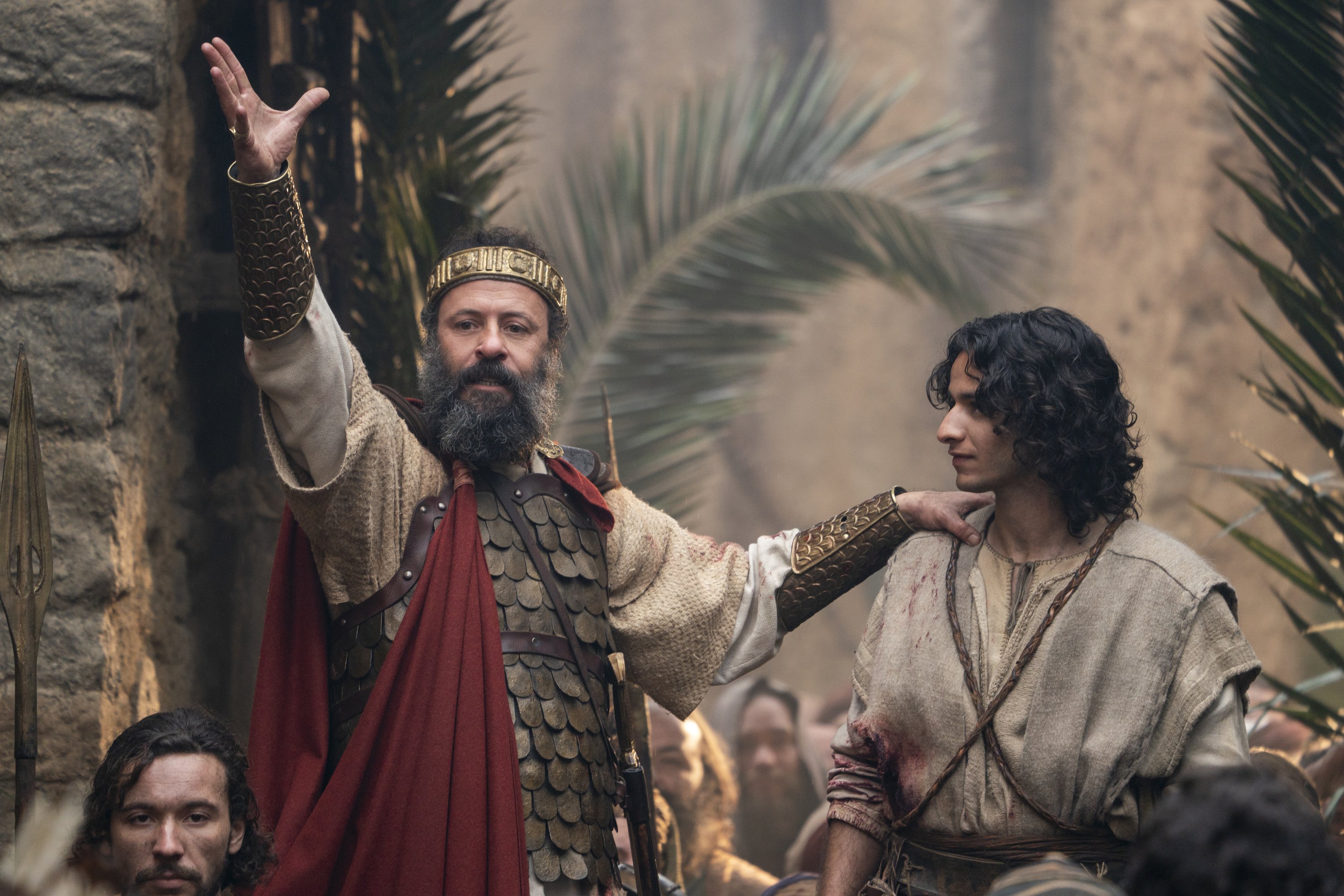 A bearded man in a gold crown, scaled armor, and red cape raises one arm while resting his hand on the shoulder of a dark-haired man in bloodstained robes, before a crowd and palm fronds.