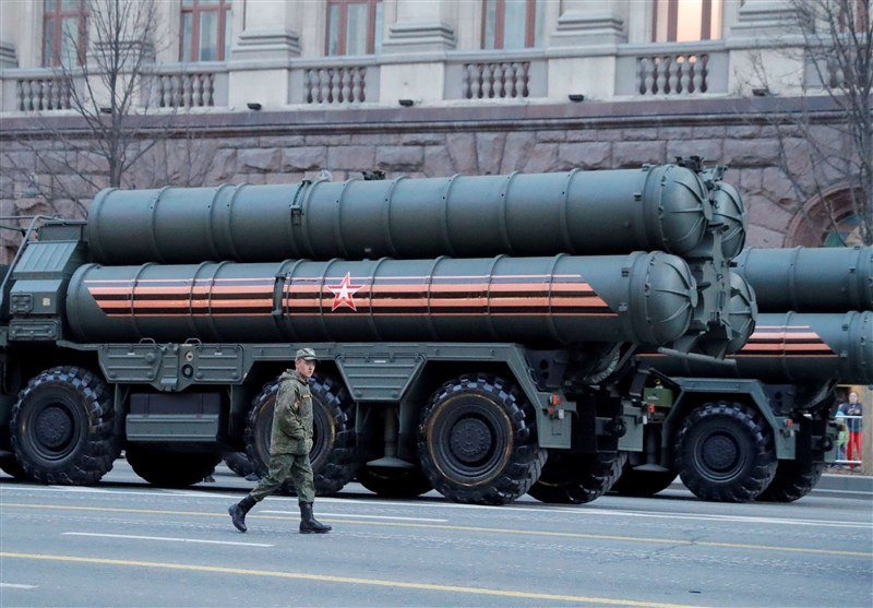 A soldier in camouflage uniform walks past a large olive-green military missile launcher truck adorned with striped ribbon and star decals on a paved street.