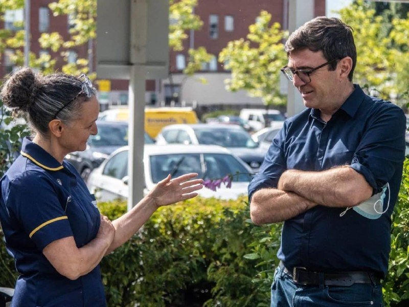 A woman in a navy uniform and a man in a blue shirt stand outdoors, conversing in front of parked vehicles and greenery.