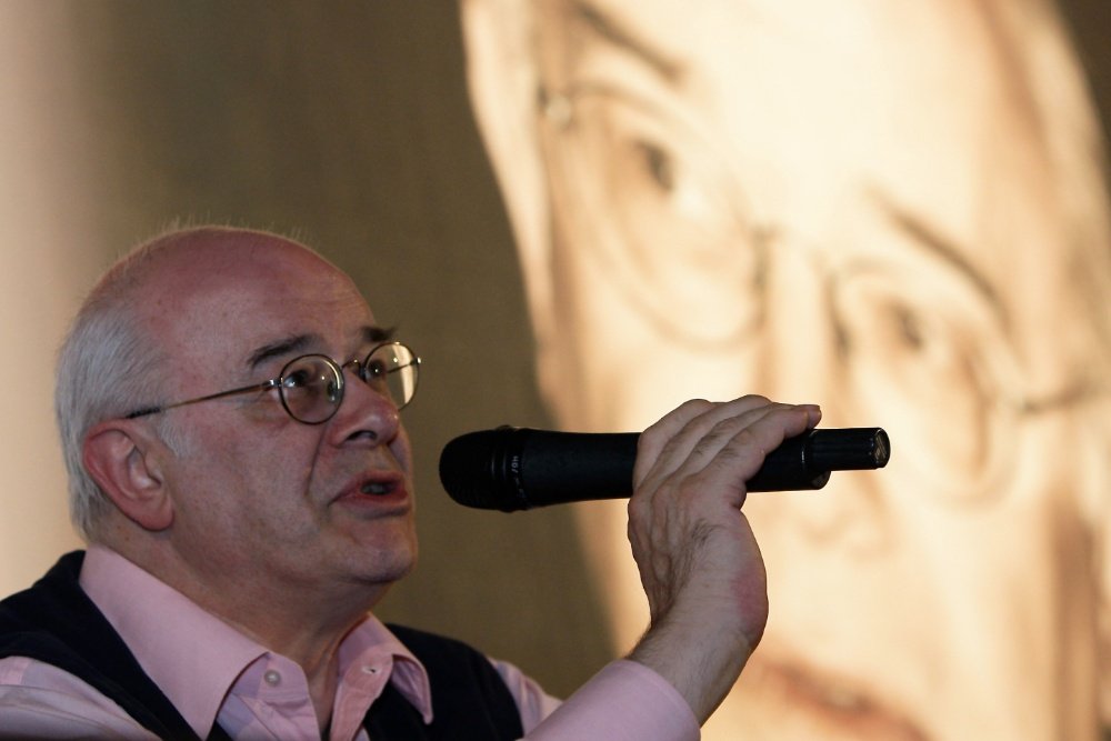 A bald man wearing round glasses and a pink shirt speaks into a microphone, with a large blurred portrait projected in the background.