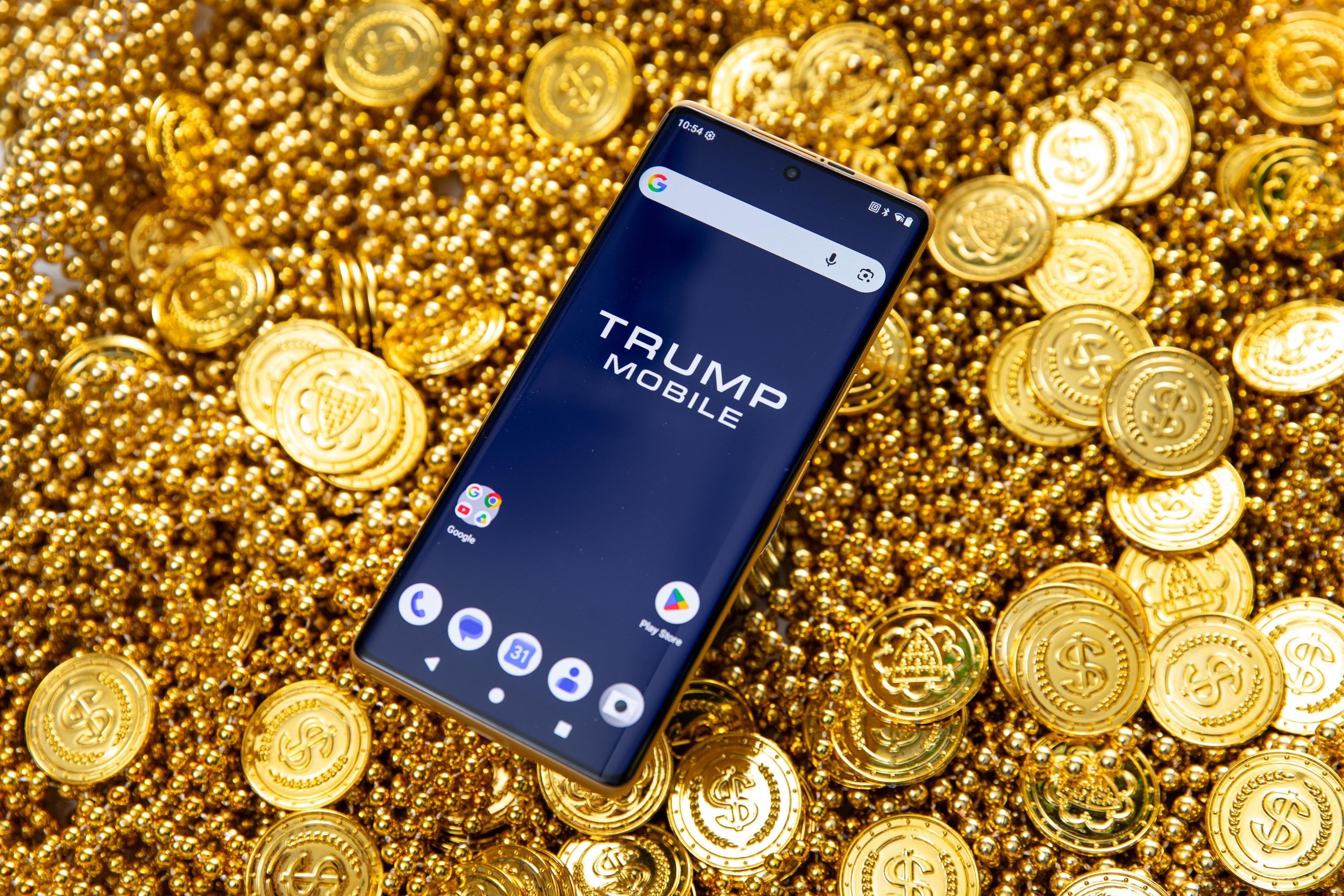 A smartphone displaying the "TRUMP MOBILE" logo on its home screen rests atop a bed of gold-colored coins and beads.