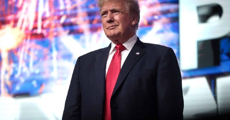 A blonde man in a dark suit and red tie stands on a stage with a large illuminated screen displaying graphics behind him.
