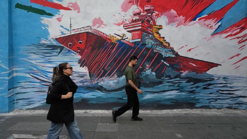 Two people walk past a street mural depicting an anthropomorphized warship with a face, painted in bold red, black, and blue brushstrokes.