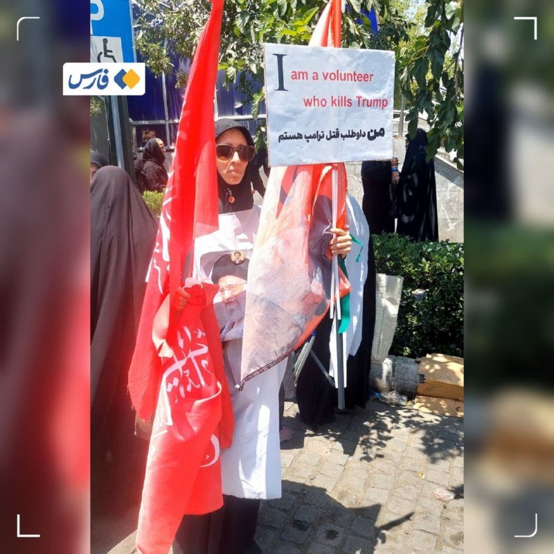 A woman in sunglasses and a headscarf holds red flags and a protest sign reading "I am a volunteer who kills Trump" with Persian text during an outdoor demonstration.
