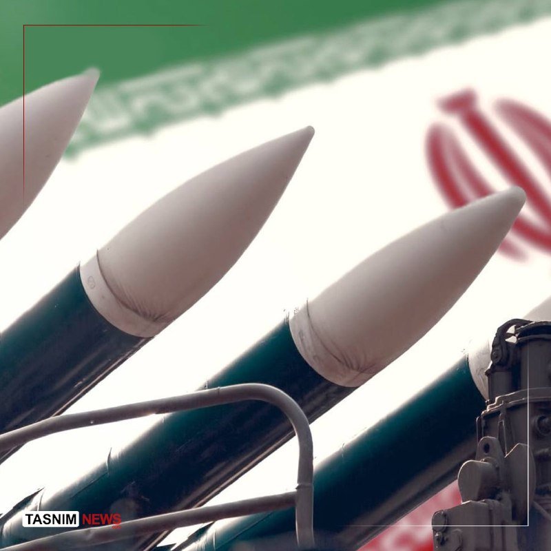 Three missiles with white nose cones and dark green bodies are mounted on a launcher, with an Iranian flag visible in the background and a "Tasnim News" watermark in the corner.