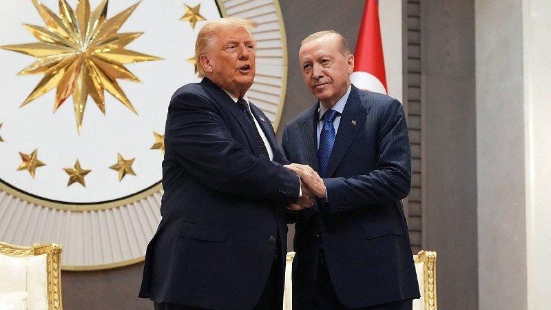 Two men in dark suits shake hands in front of a red-and-white flag and a large gold star emblem on the wall.