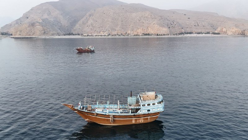 A wooden boat with an enclosed upper deck floats on calm coastal waters, with a smaller red vessel nearby and arid mountains rising in the background.