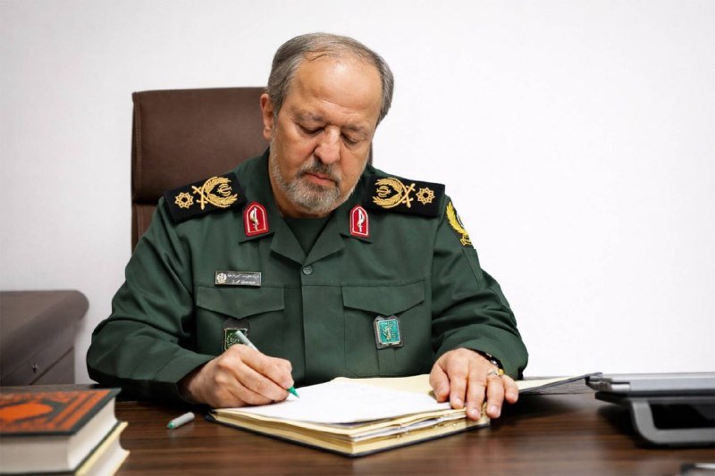 A man in an olive-green military uniform with insignia and badges writes in a notebook while seated at a desk in a plain office.