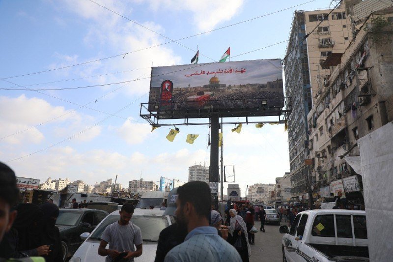 Pedestrians and vehicles move through a crowded urban street beneath a large billboard displaying Arabic text and a Palestinian flag, with power lines crossing the partly cloudy sky.
