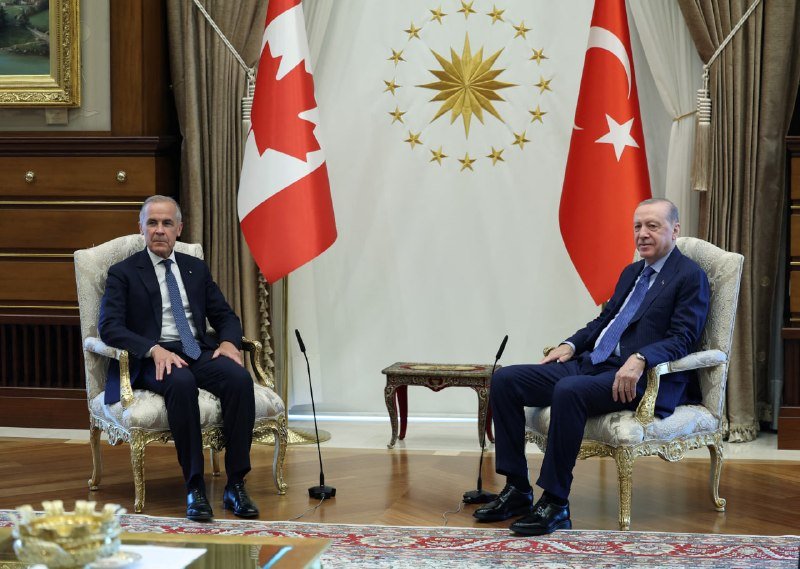 Two men in dark suits sit in ornate chairs, flanked by Canadian and Turkish flags with a gold star-and-crescent emblem on the wall behind them.