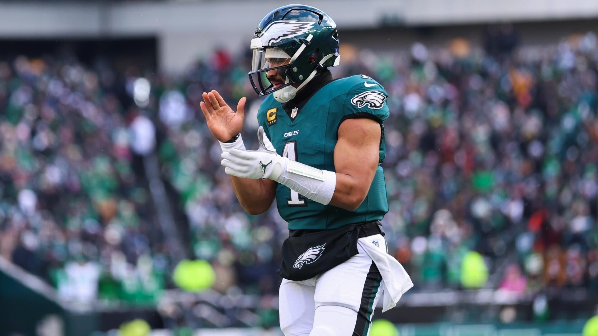 A Philadelphia Eagles football player wearing a green #1 jersey and helmet claps his hands in front of a blurred stadium crowd.