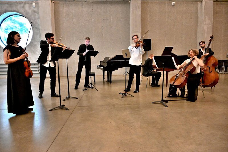 A string ensemble with a pianist performs in a spacious industrial-style room featuring concrete walls and a large illuminated blue sphere.
