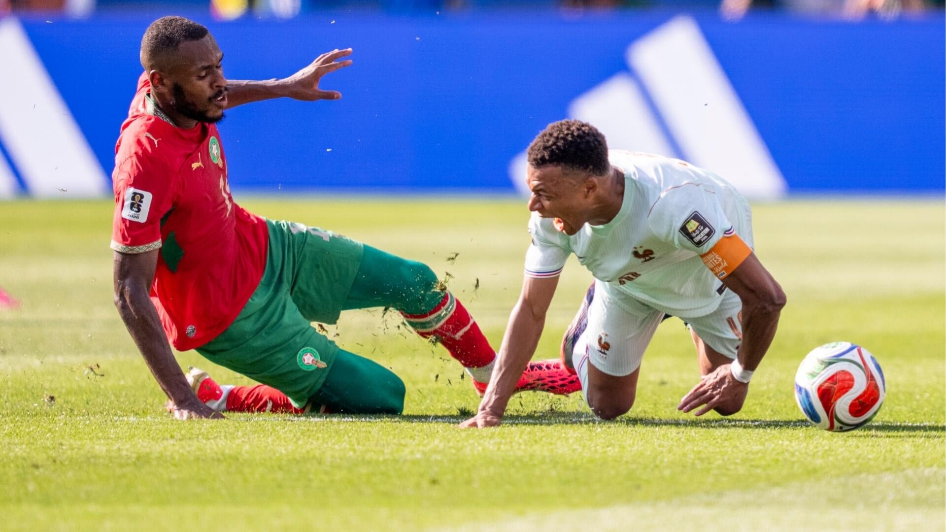 Two soccer players slide on the grass during a match, one in a red and green uniform and the other in white, competing for a colorful ball.