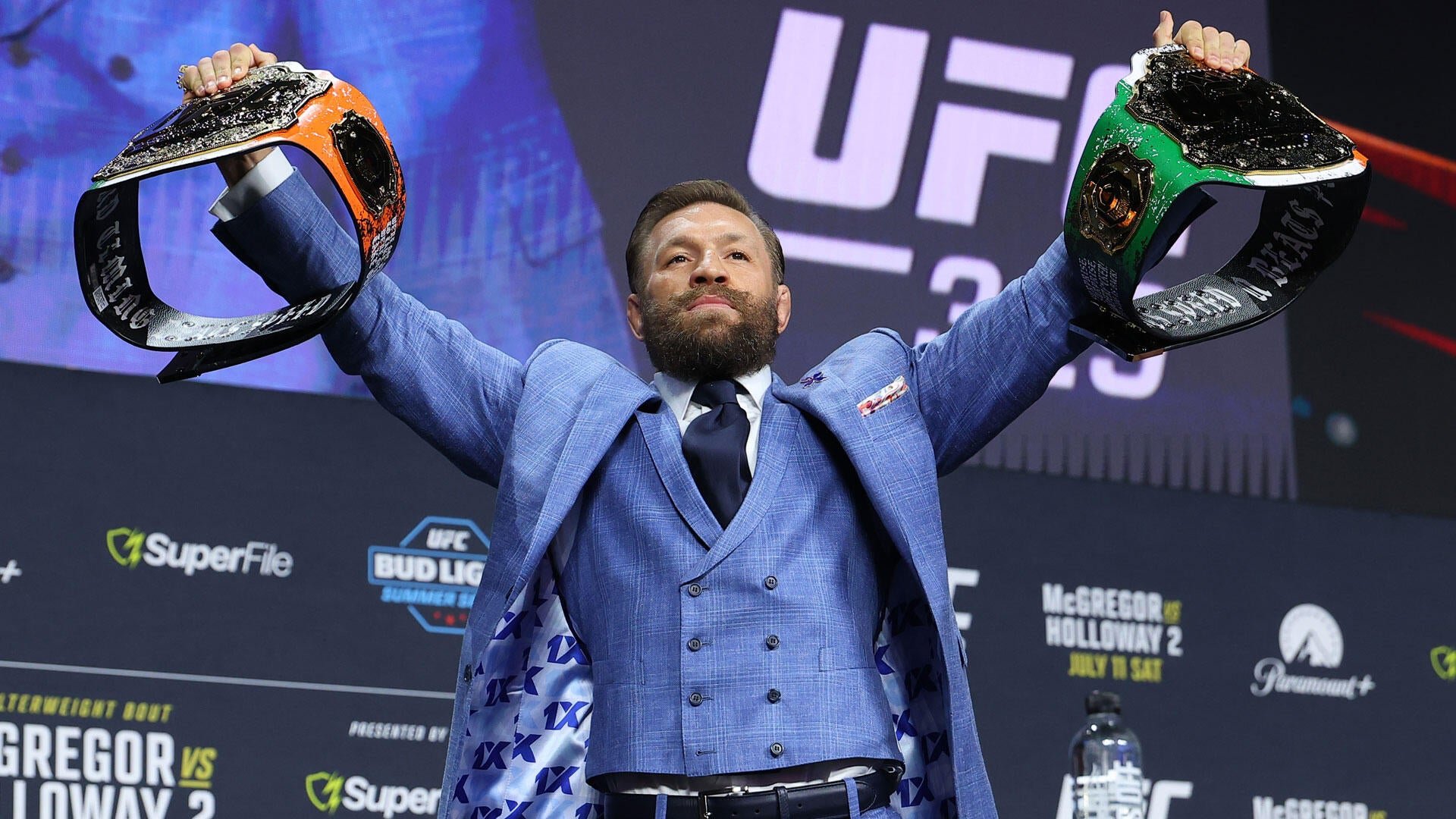 Bearded man in a blue plaid suit raises two championship belts overhead at a UFC promotional event backdrop displaying "McGregor vs Holloway 2."