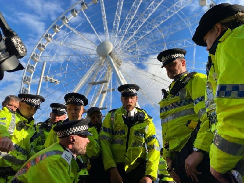 Police officers in high-visibility yellow jackets and checkered caps gather outdoors, with a large white Ferris wheel visible against a blue sky in the background.