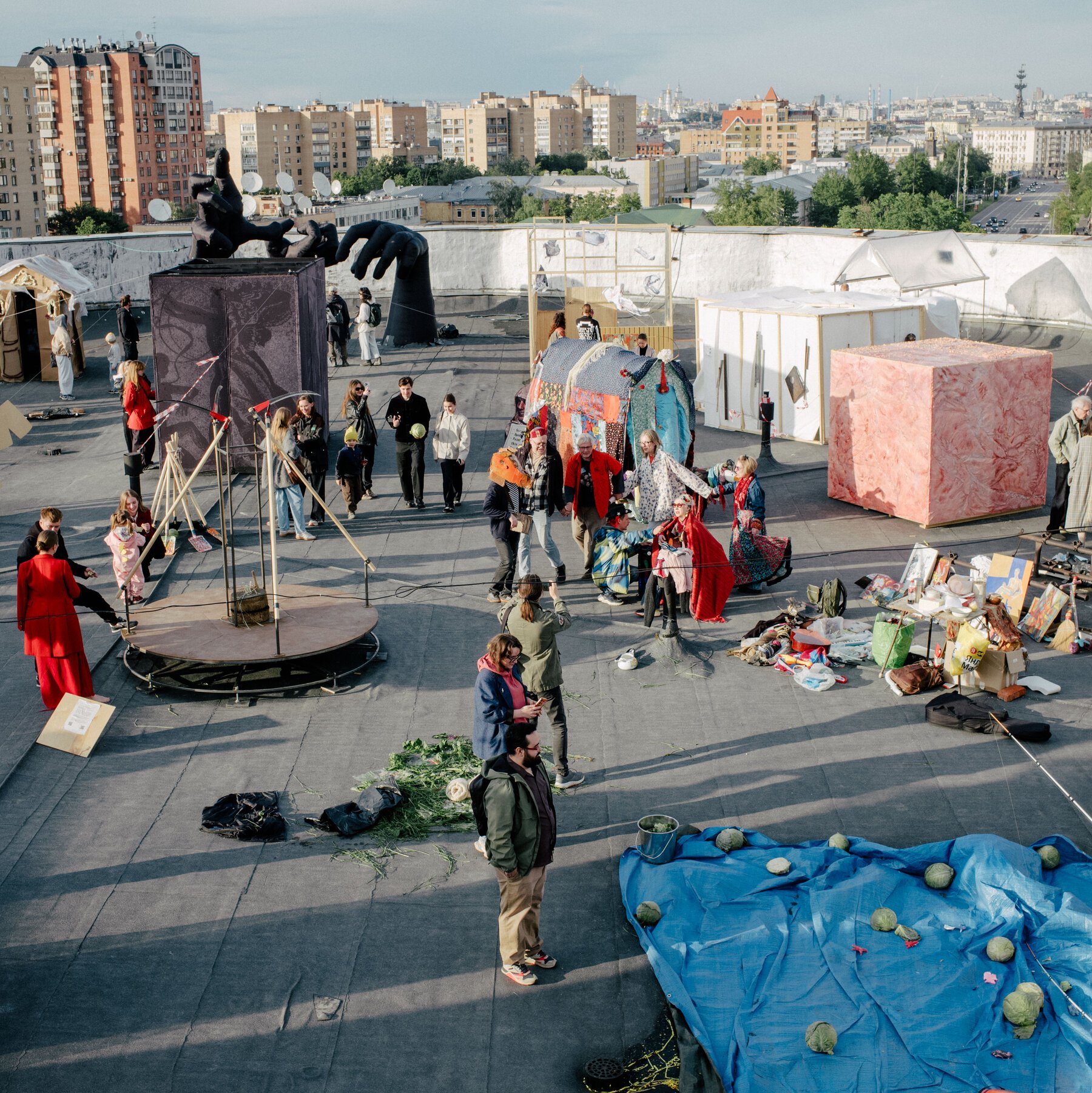 People gather on a rooftop featuring art installations, sculptures, tents, and a city skyline in the background.