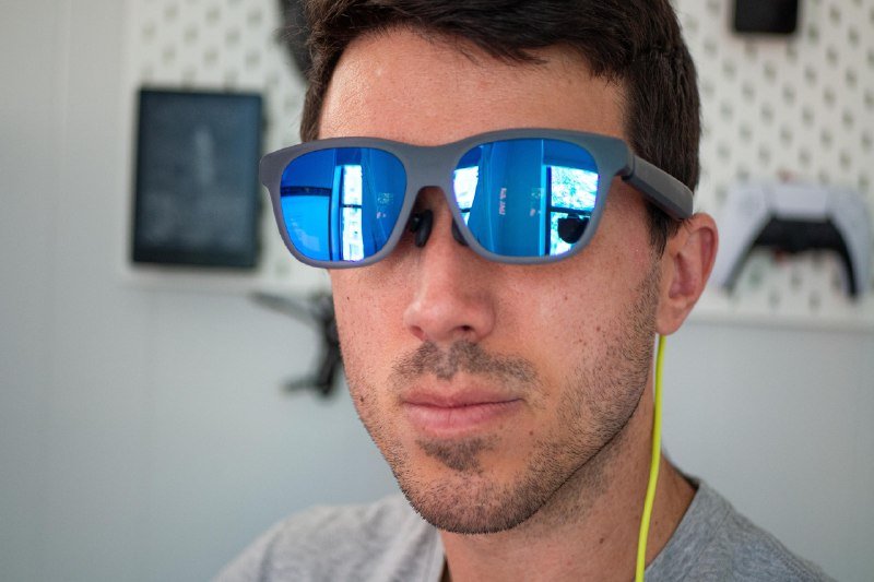 A man wearing blue-mirrored sunglasses and yellow earbuds poses in front of a pegboard wall displaying a game controller and framed item.