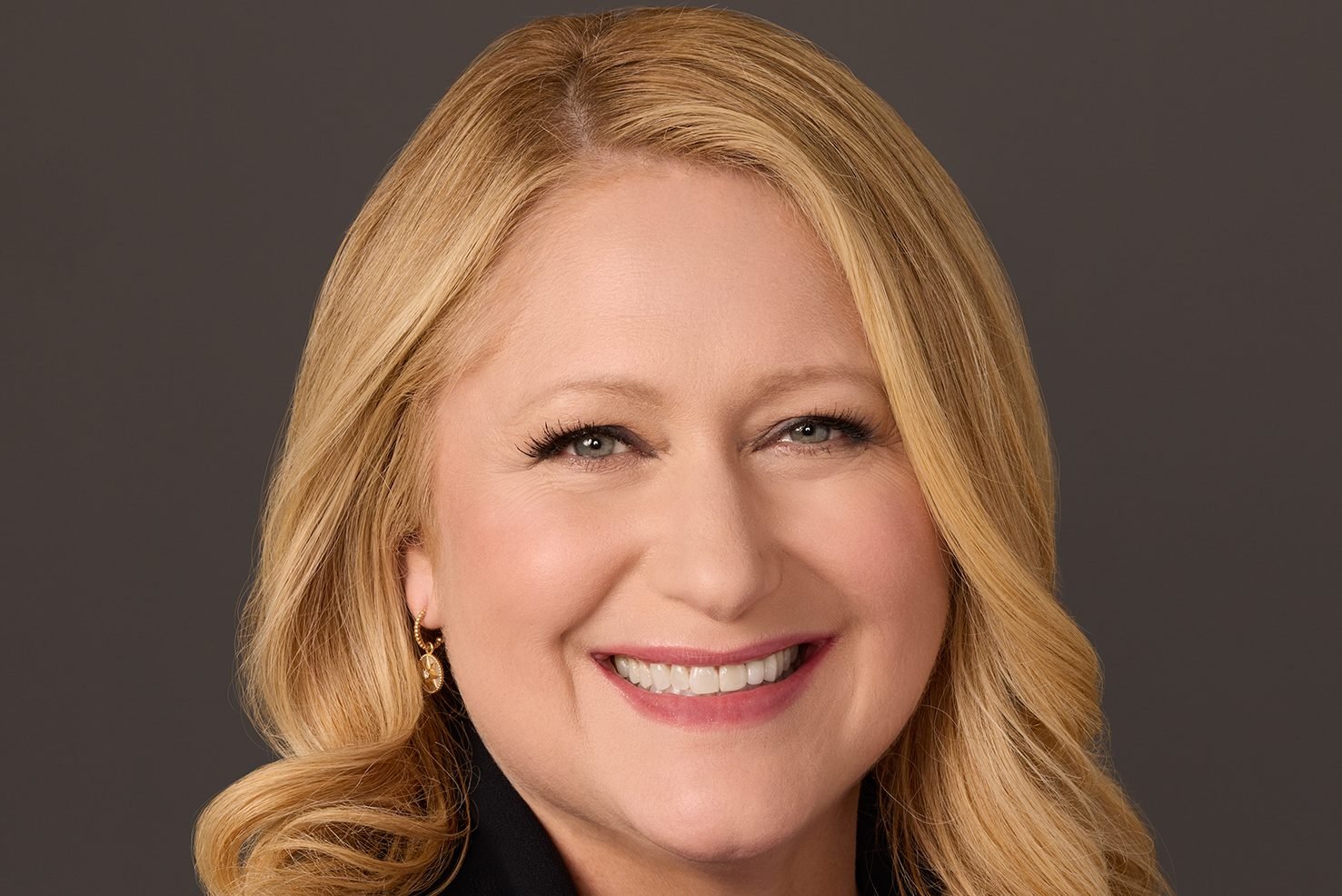 A smiling woman with long blonde hair and gold earrings is pictured against a dark brown background in a professional headshot.