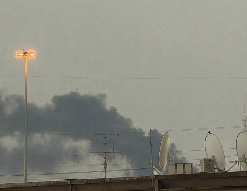 Thick dark smoke rises behind a rooftop equipped with satellite dishes, antennas, and a lit streetlamp under a hazy gray sky.