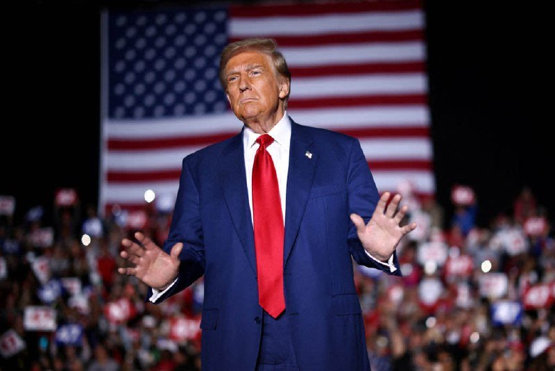 A man in a blue suit and red tie stands on stage with arms outstretched in front of a large American flag and a crowd holding campaign signs.