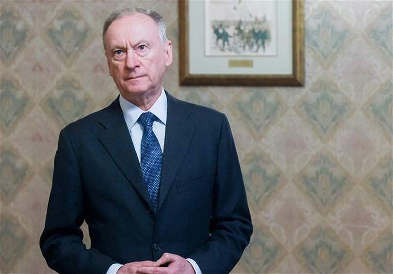 An older man in a dark suit and blue tie stands indoors against patterned wallpaper, with a framed equestrian illustration hanging behind him.