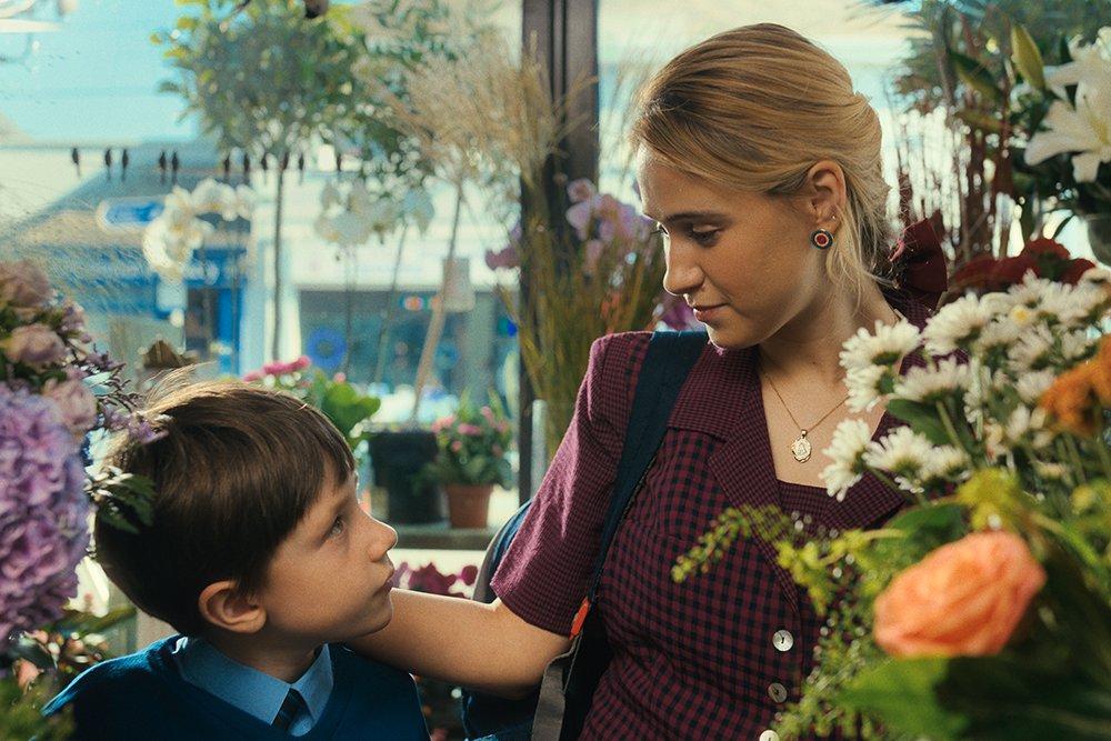 A blonde woman in a maroon checkered dress looks down at a young boy in a blue shirt and tie, surrounded by bouquets of flowers in a shop.