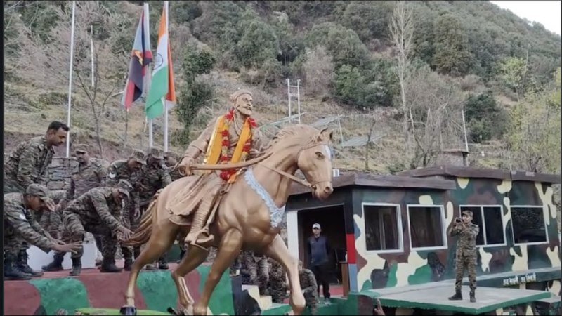 Indian Army soldiers in camouflage uniforms stand beside a golden statue of a warrior on horseback adorned with a flower garland, near flags and a camouflaged structure in a mountainous setting.