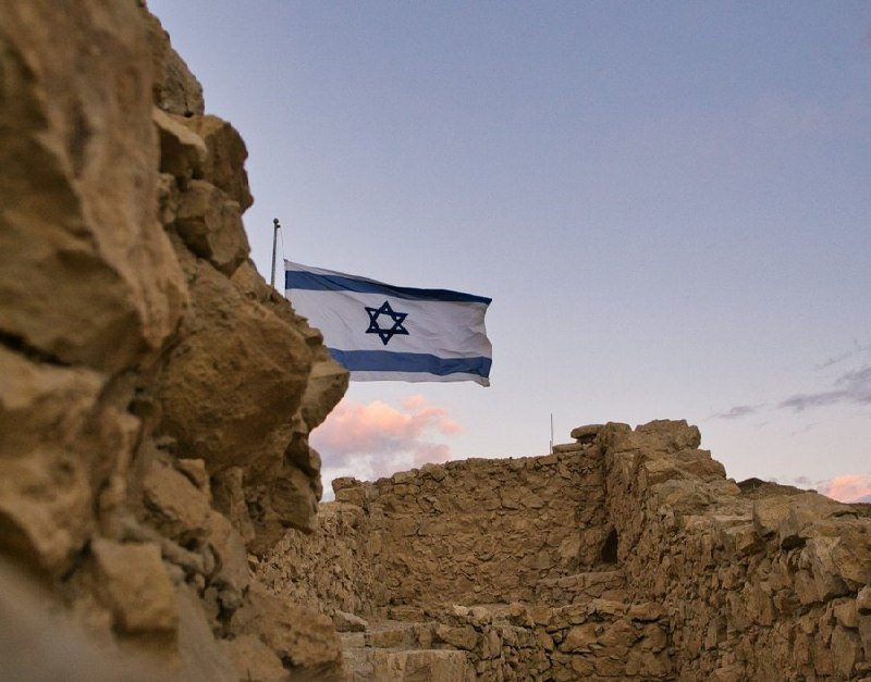 The Israeli flag flies above ancient stone ruins under a partly cloudy sky at dusk.