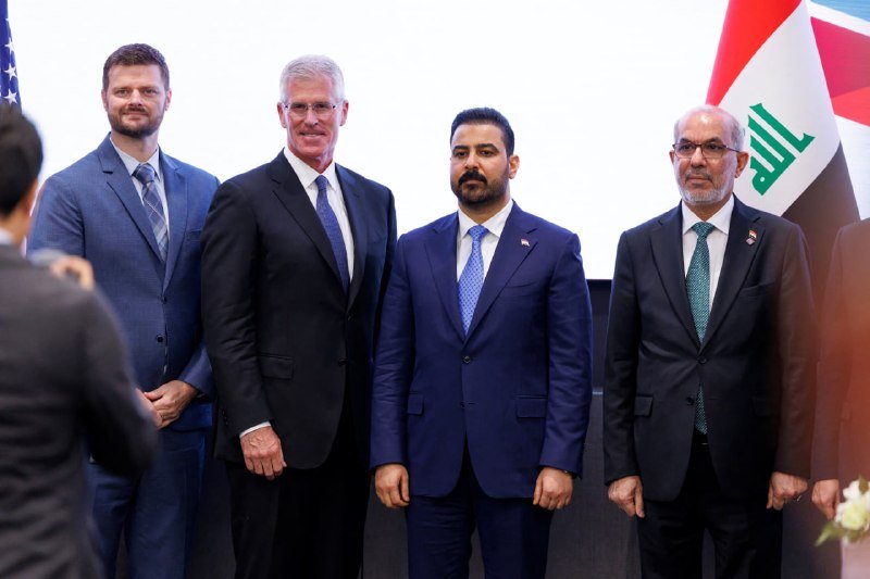 Four men in suits pose for a photo beside an American flag and an Iraqi flag at a formal event.