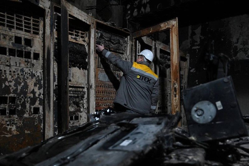 A worker in a hard hat and gray jacket labeled "ДТЕК" inspects burn-damaged electrical equipment and wiring inside a charred server or control room.
