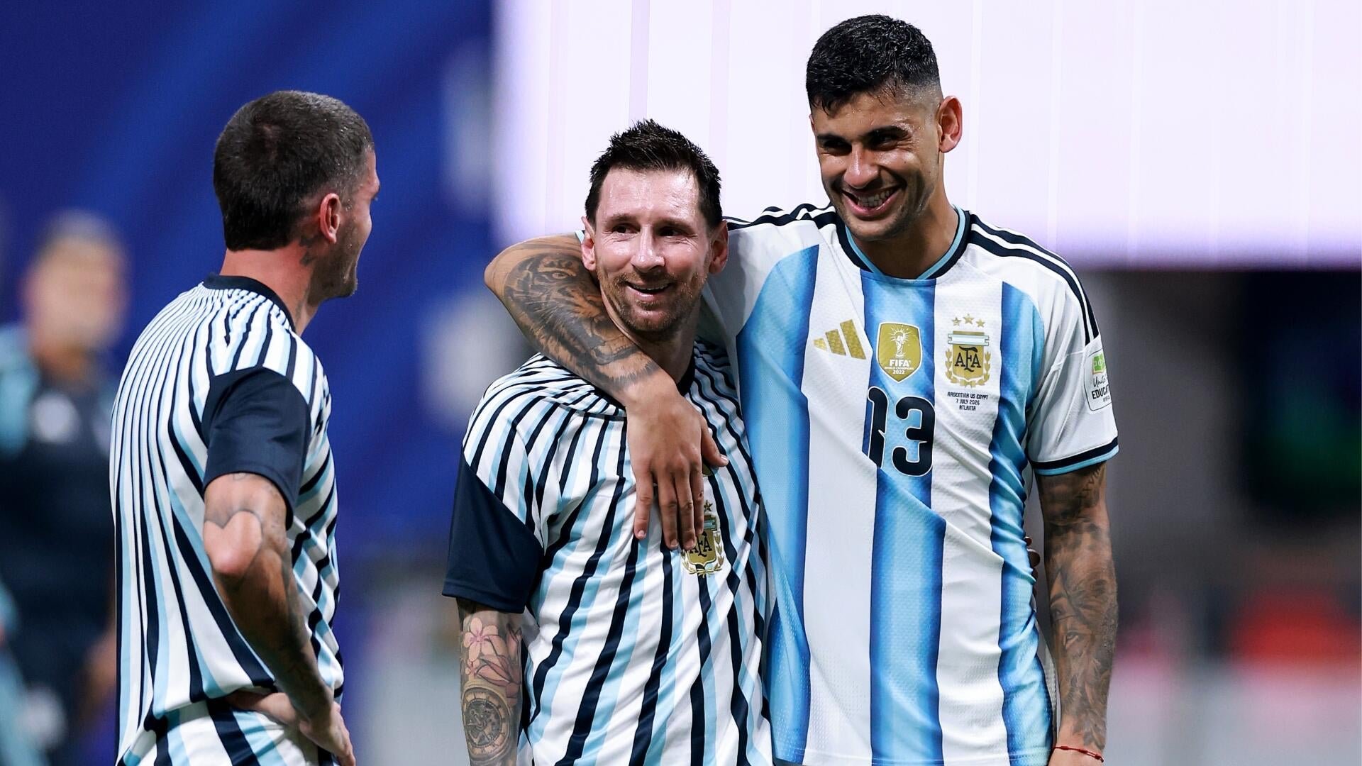 Three soccer players in Argentina jerseys smile and embrace on a field, with one wearing jersey number 13.