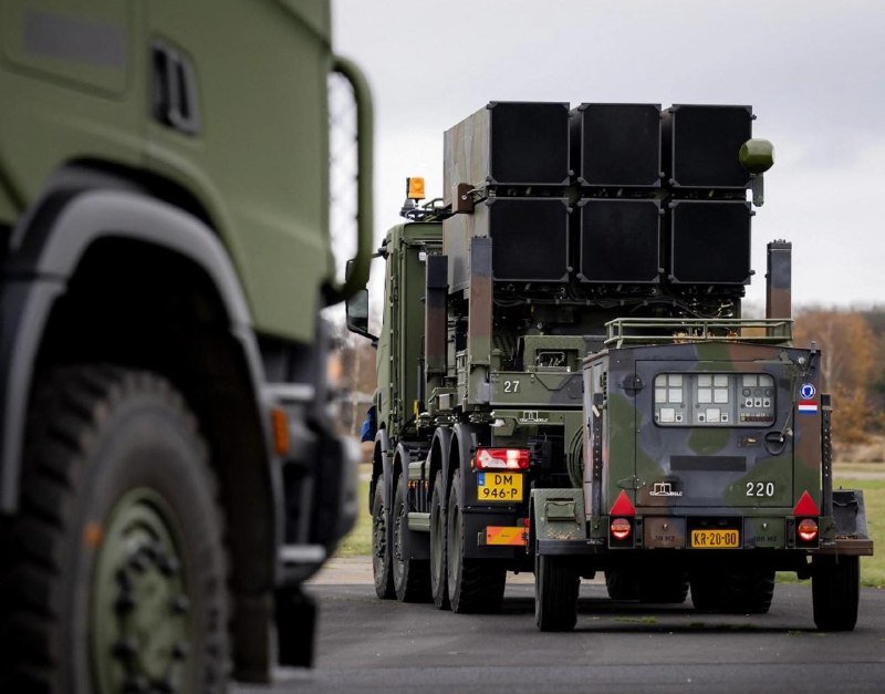 Military trucks carrying a large camouflaged missile launcher system travel along a paved road under an overcast sky.