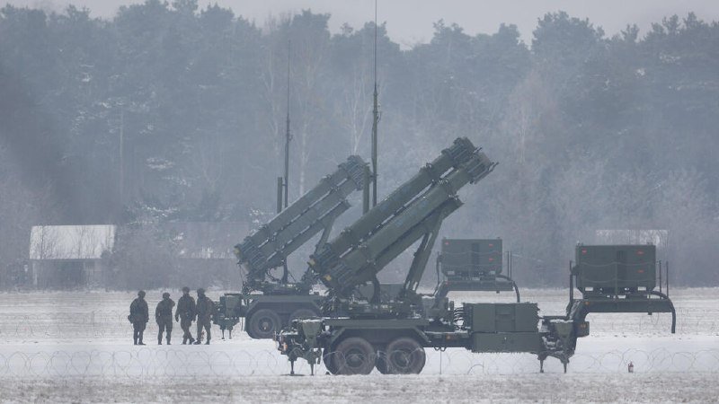 Four soldiers stand beside mobile surface-to-air missile launchers deployed on a snow-covered field with a forested backdrop.