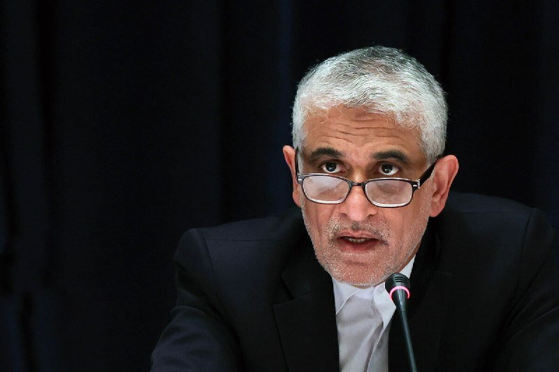 A gray-haired man with glasses speaks into a microphone against a dark background.