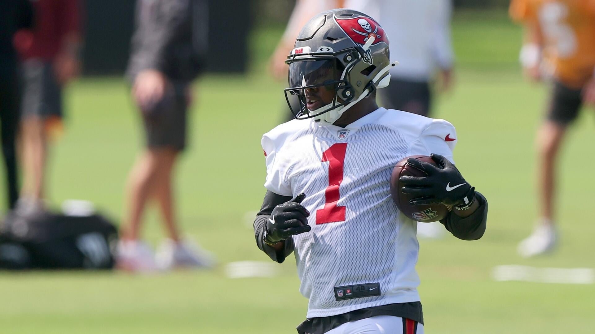 A football player wearing a white Tampa Bay Buccaneers practice jersey with the number 1 and a helmet with a dark visor carries a football on a grassy field.