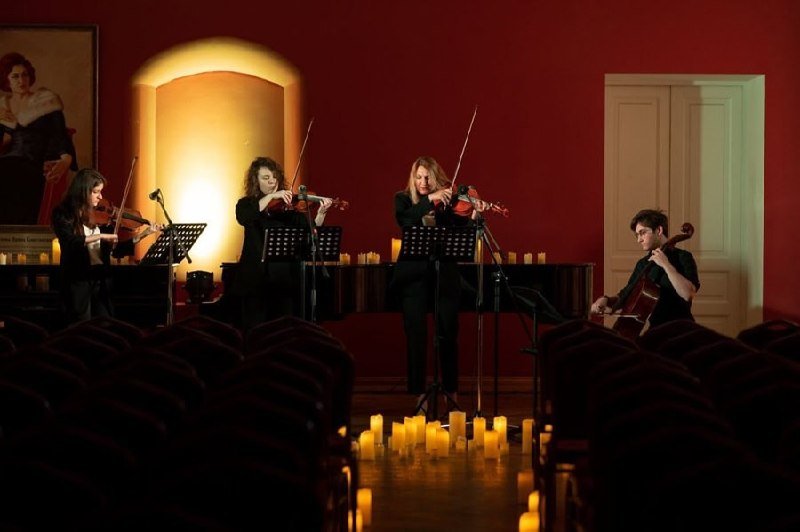 Four musicians in black play string instruments on a dimly candlelit stage with red walls and empty auditorium seats in the foreground.