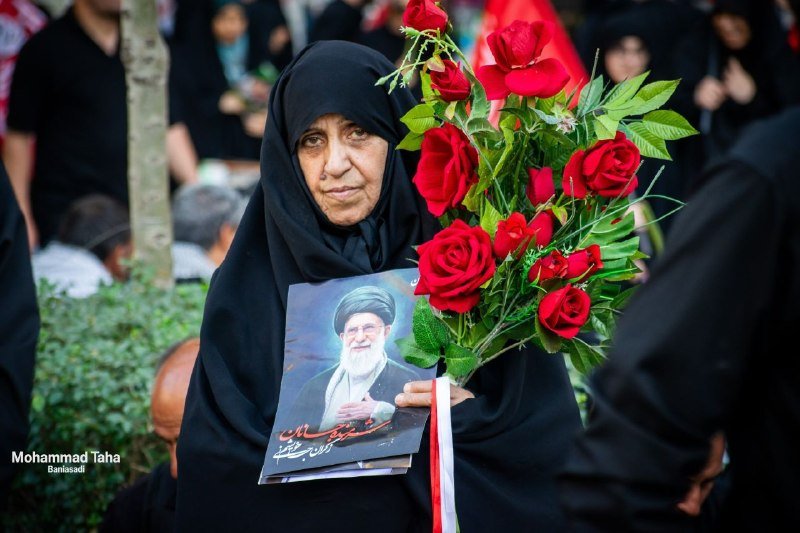 A woman in black holds a portrait of a bearded cleric in a turban and a bouquet of red roses.