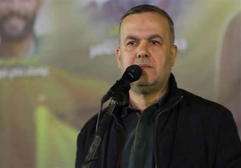 A man in a black jacket stands at a microphone, with a yellow banner featuring unreadable text visible in the background.