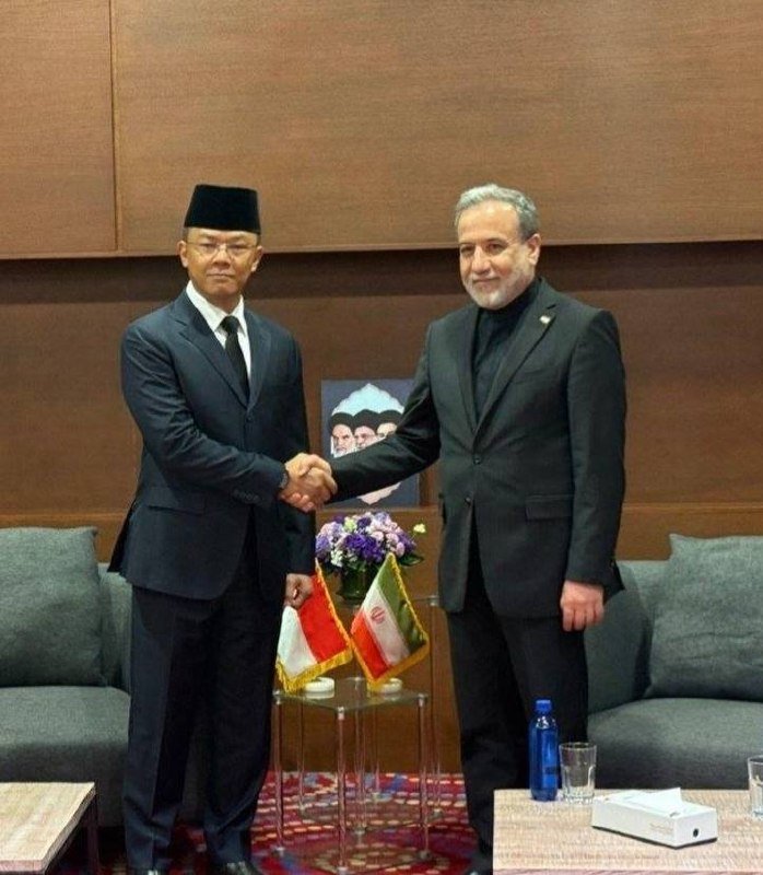 Two men in dark suits shake hands in a wood-paneled room, flanked by Indonesian and Iranian flags and a framed portrait.