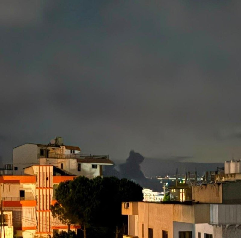 A dark plume of smoke rises above a coastal city's rooftops at dusk, with illuminated buildings and trees in the foreground.