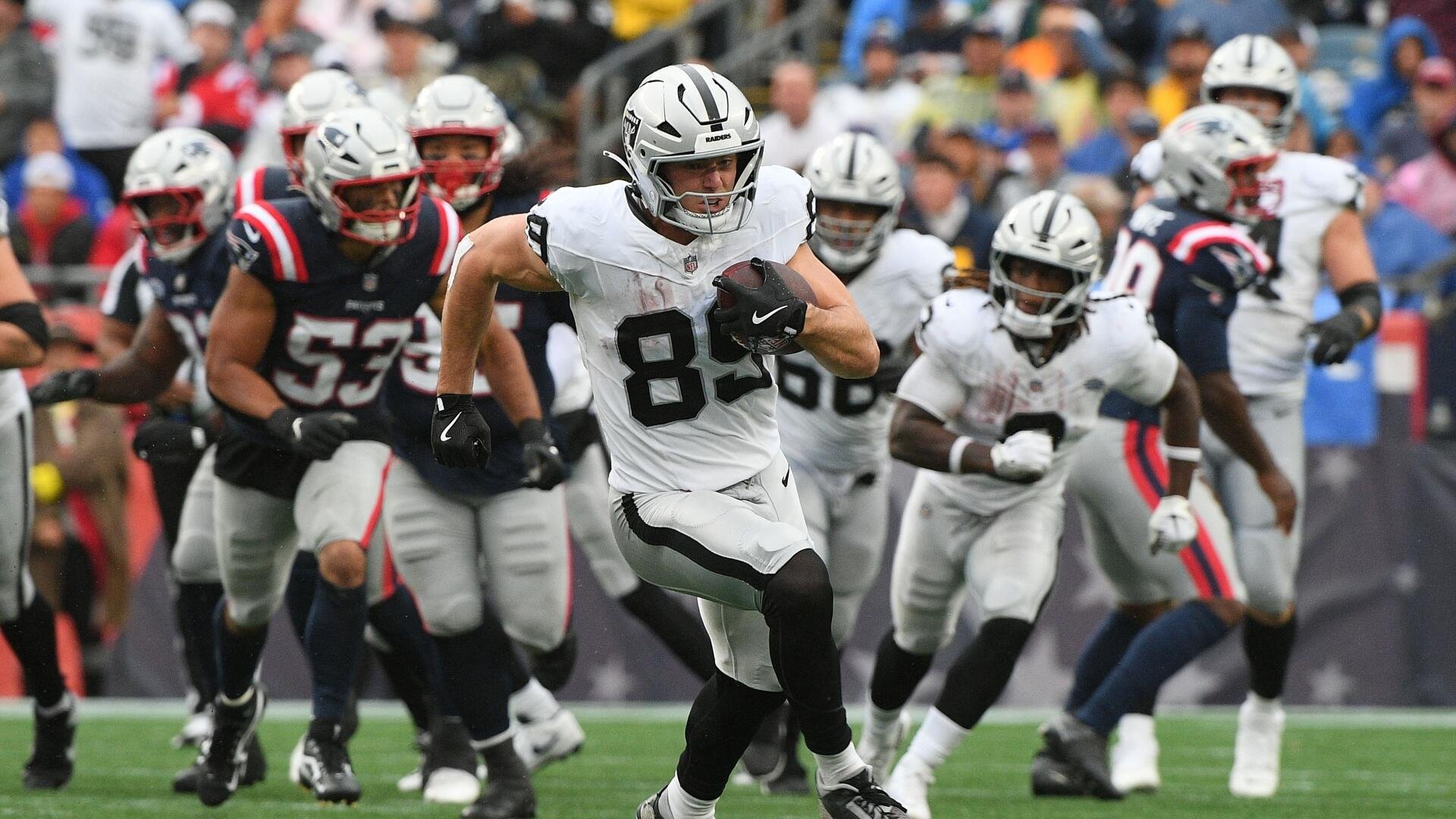 A football player wearing a white "Raiders" jersey with the number 82 runs with the ball, pursued by players in navy "Patriots" uniforms on a field before a crowd.