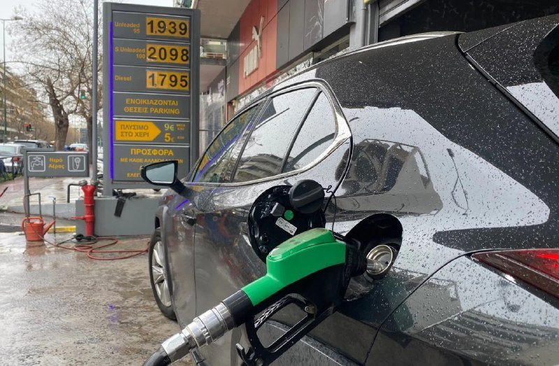 A dark-colored car is being refueled at a gas station, with a green and black nozzle inserted into its fuel tank beside a price sign displaying fuel costs.