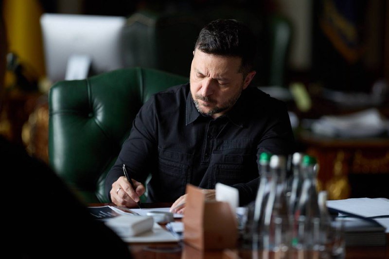 A man in a dark shirt sits in a green leather chair at a desk, writing with a pen beside several clear bottles of water.