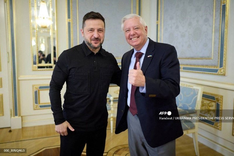 Two men pose smiling for a photo in an ornate room, one in a dark shirt and the other in a suit giving a thumbs-up.