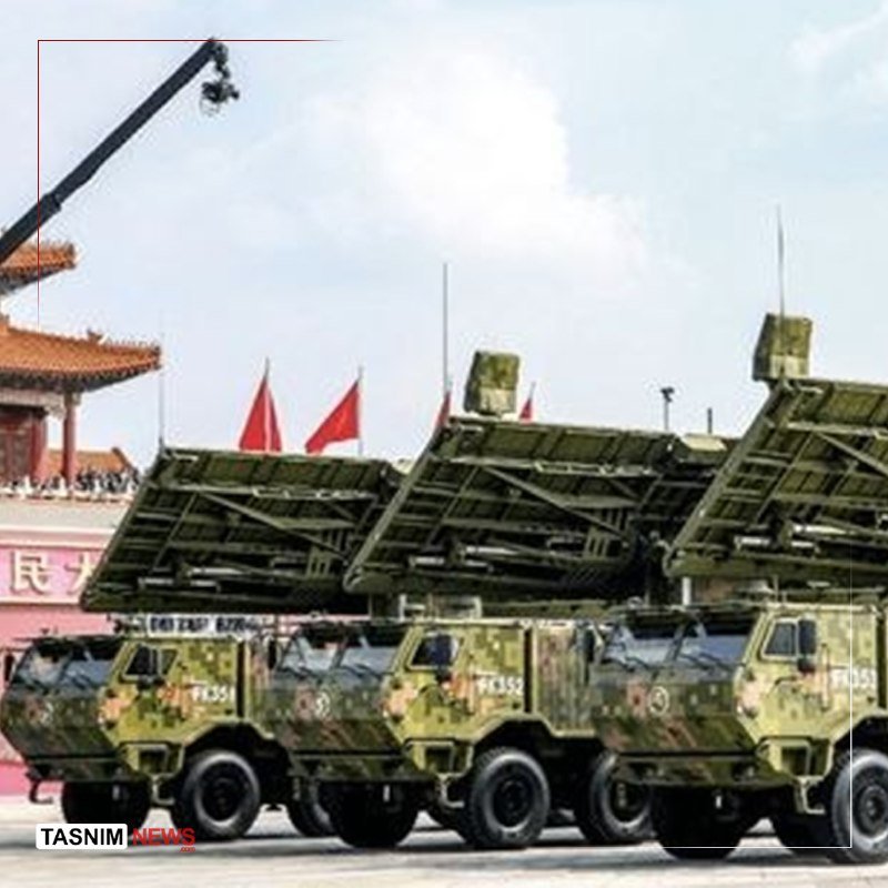 Military vehicles with camouflage paint and mounted missile launcher arrays are lined up in formation during a parade.