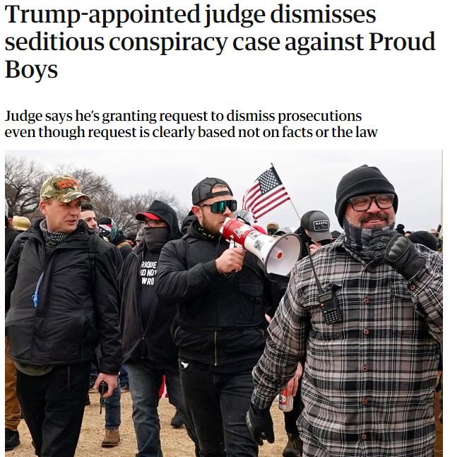 A group of people in dark winter clothing, some with face coverings, gather outdoors, with one person speaking into a red-and-white megaphone and another holding an American flag.